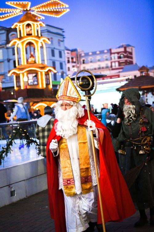 A festive scene featuring a man in a red robe and white beard, accompanied by a figure in dark clothing, amidst holiday lights.