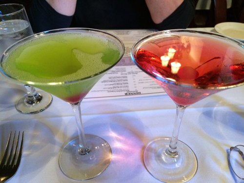 Two colorful cocktails in martini glasses: one green and bubbly, the other red with a cherry, on a white tablecloth.
