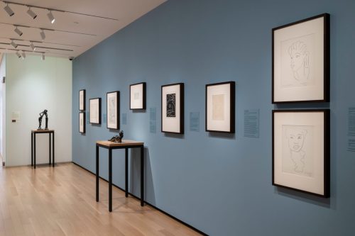 Art gallery interior featuring framed artworks on a blue wall, with sculptures displayed on small tables.