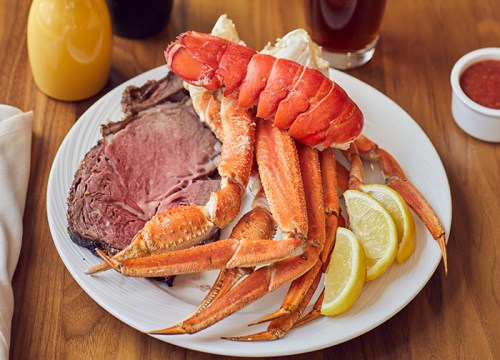 A plate featuring prime rib, lobster tail, crab legs, and lemon wedges, served with a side of dipping sauce.