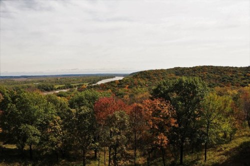 A scenic view of a river winding through colorful autumn trees and rolling hills under a cloudy sky.