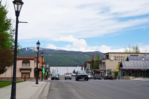 A scenic view of a lakeside town with mountains in the background, featuring shops and parked cars along the street.