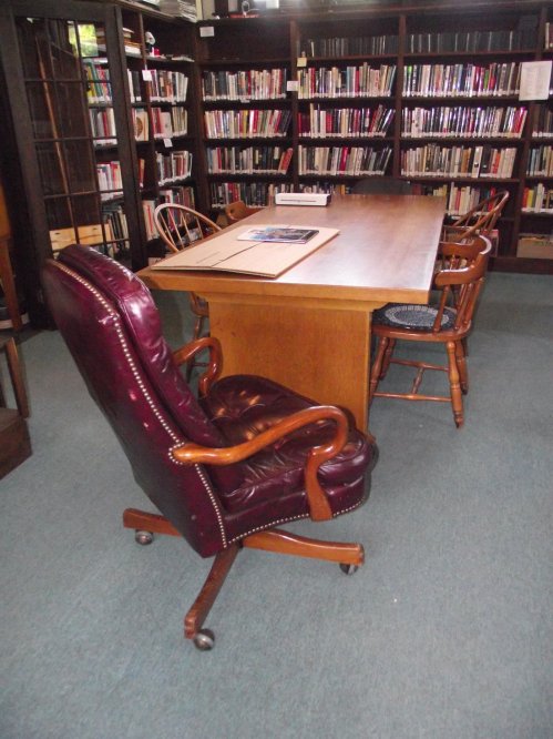 A wooden table surrounded by chairs in a library, with bookshelves filled with books in the background.