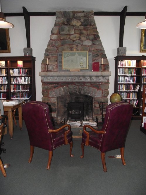 Cozy library interior featuring two chairs facing a stone fireplace, surrounded by bookshelves filled with books.