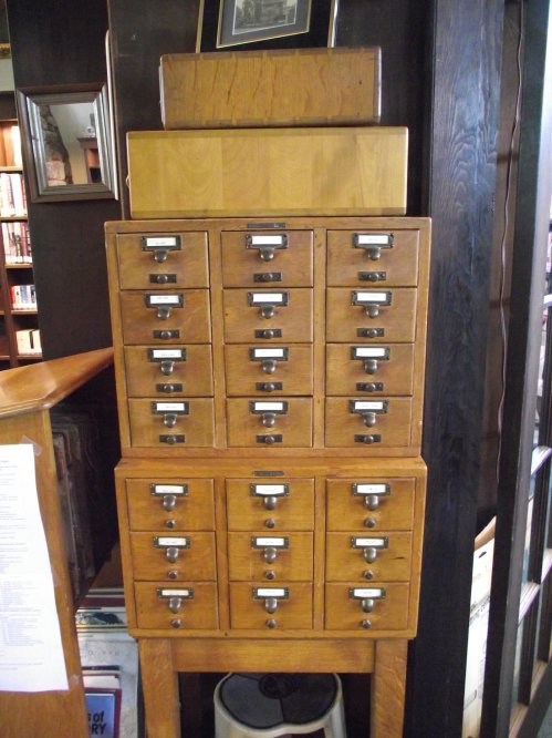 A tall wooden card catalog with multiple drawers, stacked on a wooden base, in a library setting.