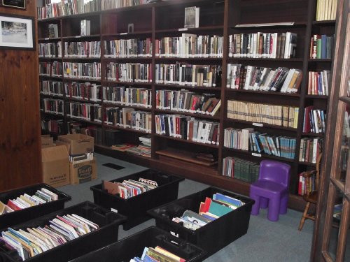 A cozy library with shelves of books, boxes of books on the floor, and a small purple chair in the corner.