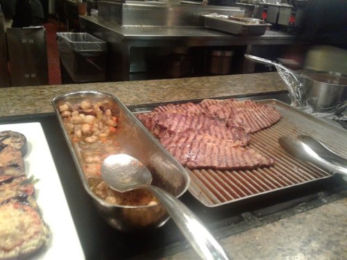 A buffet setup featuring grilled steak slices and a dish of mixed vegetables in a metal serving tray.