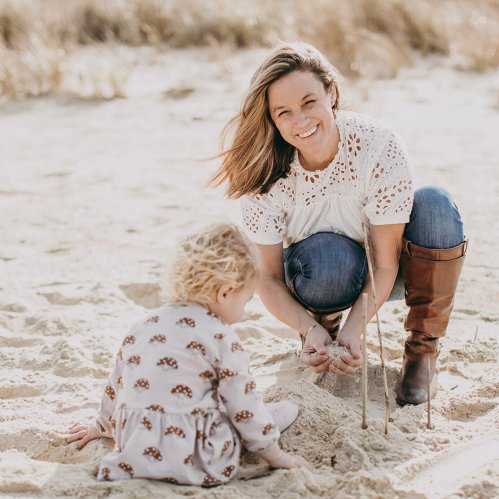 A woman smiles while playing in the sand with a young child on a beach.