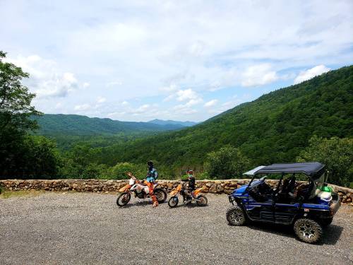 Two riders on dirt bikes and a side-by-side vehicle parked near a scenic overlook of green mountains and blue sky.