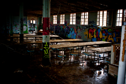 Abandoned room with graffiti-covered walls, long tables, and puddles on the floor, illuminated by dim light.