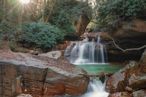 A serene waterfall cascades over rocks, surrounded by lush greenery and sunlight filtering through the trees.
