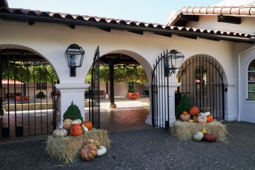 A courtyard entrance with arched gates, flanked by hay bales and pumpkins, under a clear blue sky.