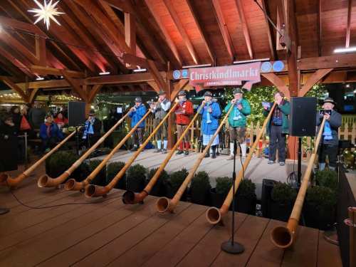A group of musicians in colorful outfits play large wooden horns on a festive stage at a Christmas market.