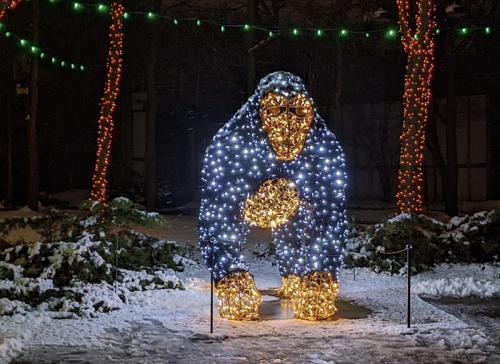 A large gorilla sculpture made of lights stands in a snowy area, surrounded by festive holiday decorations.