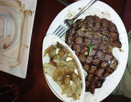 A grilled steak on a plate with a side of seasoned potatoes and herbs, served on a wooden table.