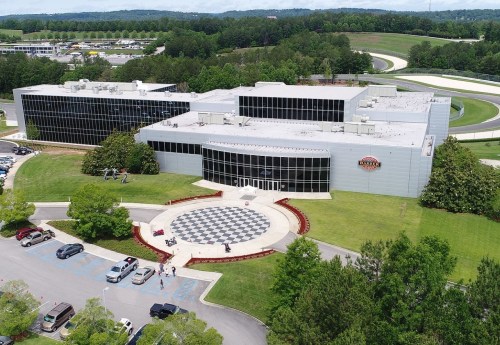 Aerial view of a modern building with a checkered pattern entrance, surrounded by greenery and parked cars.