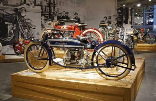 A vintage motorcycle displayed on a wooden platform, with historical photos and other motorcycles in the background.