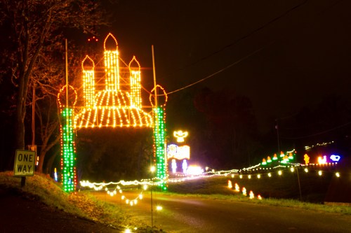 Colorful holiday lights illuminate a road, featuring a large arch and various festive decorations in the background.