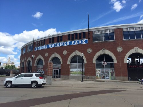 Exterior view of Modern Woodmen Park, a brick stadium with large windows and a blue sign, under a partly cloudy sky.