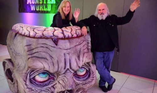 A woman and a man pose beside a large, artistic sculpture of a head with a brain, both smiling and waving.