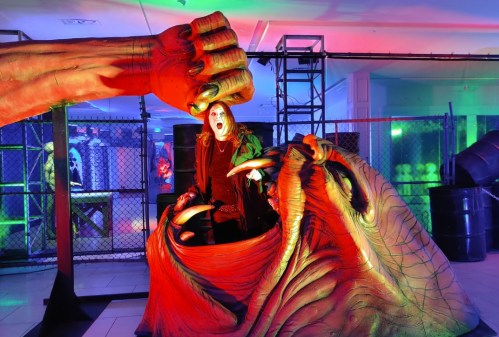 A person in dark clothing stands in a colorful, eerie setting, surrounded by large, monstrous hands and vibrant lighting.