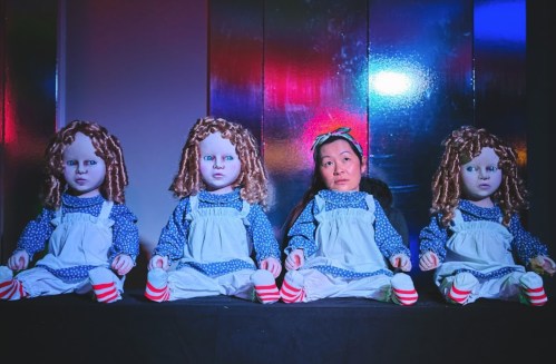 A person sits among three lifelike dolls, all dressed in blue and white outfits, against a colorful backdrop.
