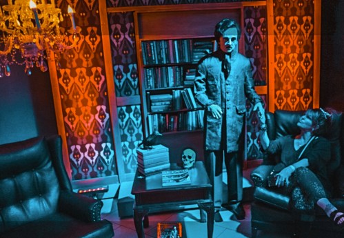 A dimly lit room with a skeleton figure, a woman in a sparkly outfit, and a bookshelf, creating a spooky atmosphere.