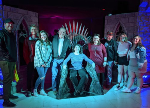 A group of eight people poses together, with one person seated on a replica throne, in a themed setting.