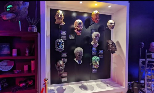 A display of various masks, including alien and monster designs, arranged on a black wall with colorful lighting.