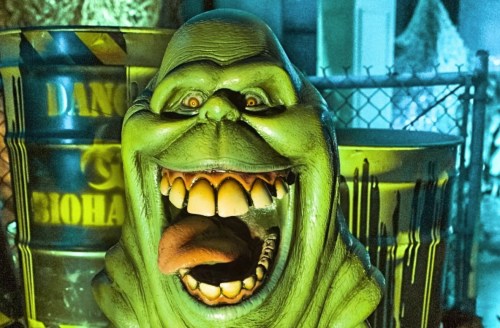 A green, ghostly figure with a wide grin and protruding tongue, set against a backdrop of barrels and eerie lighting.