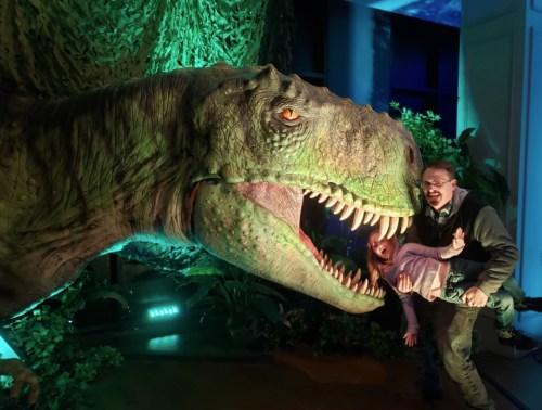 A man playfully holds a child near a large dinosaur model, with vibrant green lighting in the background.