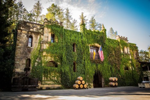 A historic stone building covered in green ivy, with an American flag and wine barrels in front, surrounded by trees.