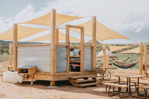 A modern wooden structure with metal siding, shaded by large fabric canopies, surrounded by hammocks and picnic tables.