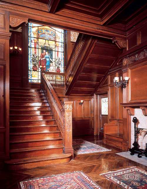 A grand wooden staircase with intricate carvings, stained glass window, and elegant decor in a historic interior.