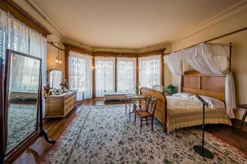 A vintage bedroom featuring a wooden bed, dresser, mirror, and large windows with sheer curtains, all on a floral rug.