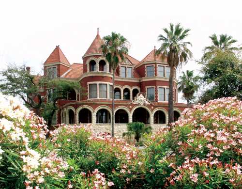 A grand red mansion with turrets, surrounded by blooming flowers and palm trees under a cloudy sky.