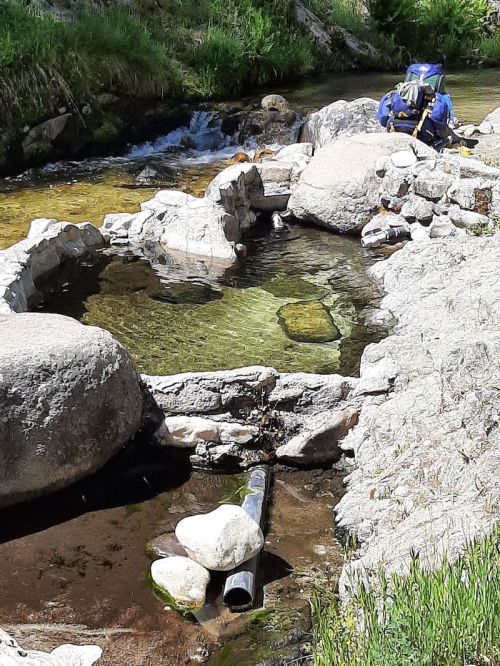 A natural hot spring surrounded by rocks, with a small stream flowing nearby and a backpack resting on the ground.