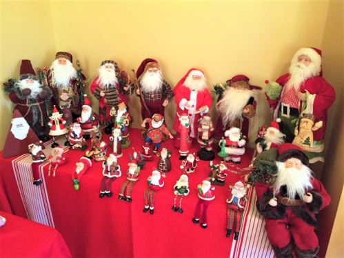 A collection of various Santa Claus figurines displayed on a red tablecloth against a yellow wall.