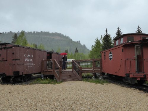Two vintage train cars are positioned near a wooden deck, with a person holding an umbrella in the background on a rainy day.