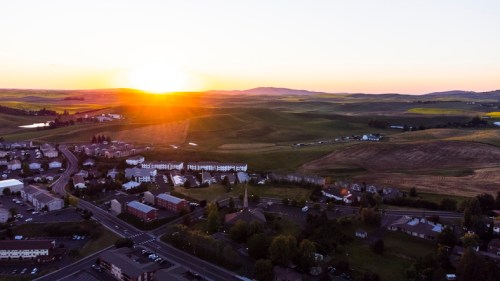 Aerial view of a sunset over rolling hills and a small town, with fields and buildings in the foreground.