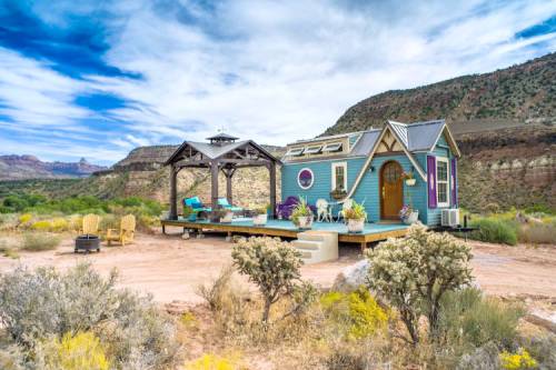 A colorful tiny house with a deck and gazebo, surrounded by desert landscape and mountains under a cloudy sky.