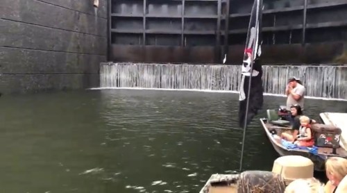 A boat with people near a waterfall in a canal, featuring a pirate flag and a stone wall backdrop.
