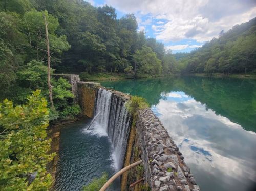 A serene waterfall cascades over a stone dam into a calm, reflective lake surrounded by lush green trees.