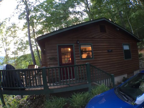 A cozy wooden cabin surrounded by trees, featuring a deck and a parked blue car in the foreground.