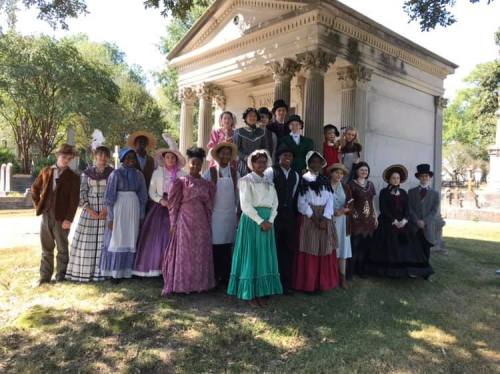 A group of people in historical costumes poses in front of a monument in a cemetery, surrounded by trees.