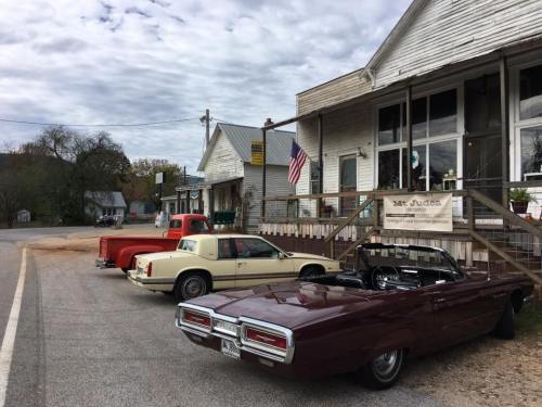 A vintage car lineup in front of a rustic building with an American flag, set in a small town.