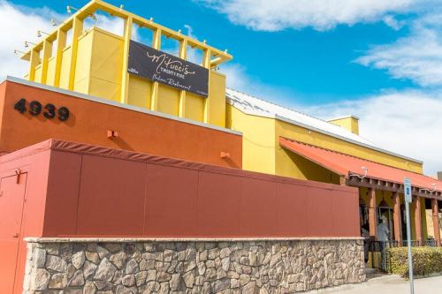 Exterior of a colorful restaurant with a stone wall, featuring a sign that reads "M'Vine's" under a blue sky.