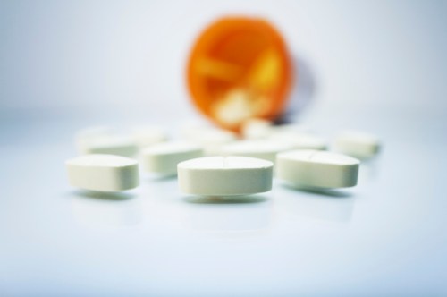 A close-up of white pills scattered on a surface, with a tipped-over orange prescription bottle in the background.