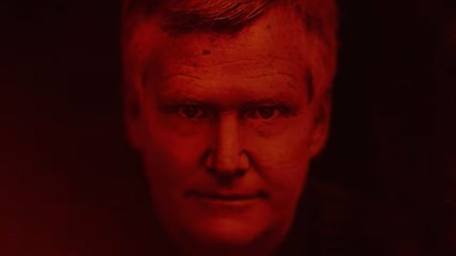 A close-up portrait of a man with short hair, illuminated in red light, creating a dramatic and intense atmosphere.