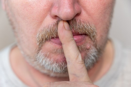 A close-up of a man with a mustache and beard, holding a finger to his lips in a shushing gesture.